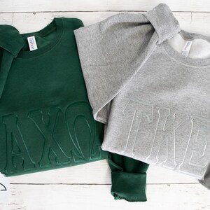 May include: Two sweatshirts with embroidered Greek letters. The sweatshirt on the left is dark green with the letters "AXO" in white. The sweatshirt on the right is gray with the letters "TKE" in white.