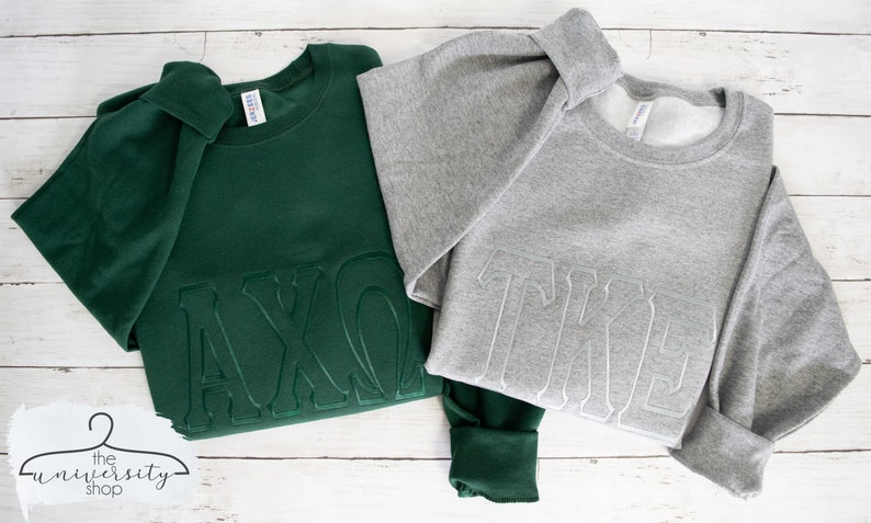 May include: Two sweatshirts with embroidered Greek letters. The sweatshirt on the left is dark green with the letters "AXO" in white. The sweatshirt on the right is gray with the letters "TKE" in white.
