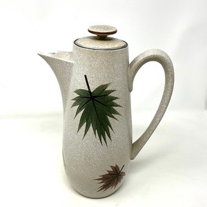 May include: A white ceramic coffee pot with a brown lid and green and brown leaf designs.