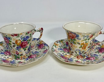 May include: Two floral teacups and saucers with a white background and a colorful floral pattern. The cups have gold rims and the saucers have a scalloped edge.