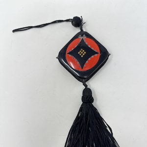 May include: A black square-shaped pendant with a red and black circle design. The circle has a black diamond shape with gold accents. The pendant has a black tassel.