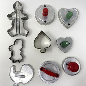 May include: A set of nine vintage metal cookie cutters with red and green handles. The cutters are shaped like a gingerbread person, a girl, a turkey, a heart, a spade, and three round shapes.