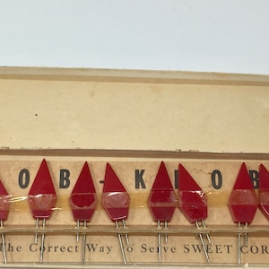 May include: A box of red plastic corn holders with the words "The Correct Way to Serve Sweet Corn" printed on the front. The holders are shaped like triangles and have a small metal spike on the bottom.