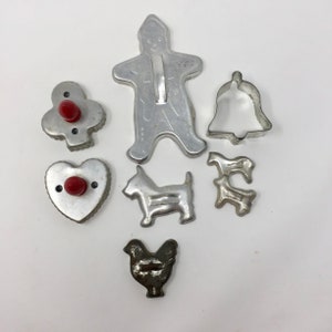 May include: A set of vintage metal cookie cutters, including a gingerbread man, a bell, a heart, a clover, a chicken, and two dogs. The cutters have red plastic handles.