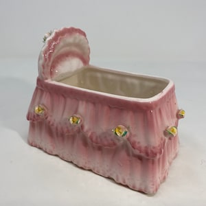 May include: A pink ceramic baby crib planter with a white ruffled canopy and yellow flower accents.