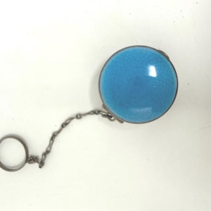 May include: A round, light blue object with a silver-colored metal rim and a matching chain. The chain connects to a small ring. The object has a glossy, smooth surface.