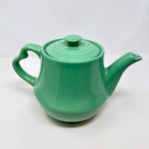 May include: A mint green ceramic teapot with a glossy finish and a visible crackle glaze pattern. The teapot features a rounded body, a curved handle, a matching lid with a small knob, and a tapered spout with a slight chip at the tip.