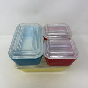May include: A set of four rectangular glass storage containers with lids. One large yellow container, one medium blue container, and two smaller red containers. Each container features a clear glass lid with a ribbed, linear textured design.
