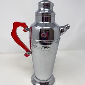 May include: A chrome cocktail shaker with a red handle. The shaker has a hammered texture on the lower portion and a spout. The handle is a bright red, and the top is a shiny chrome finish. The shaker is designed for mixing drinks.