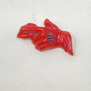May include: A red hand-shaped figurine with three black lines on the palm. The figurine is a solid color and appears to be made of a hard material. The hand is in a pointing gesture.