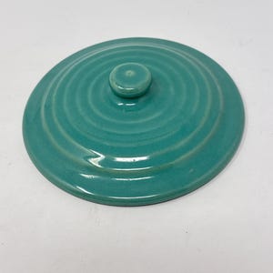 May include: A round, teal-colored ceramic lid with a glossy finish and concentric circular ridges radiating from a small, centered knob handle. The lid features a smooth, glazed surface and a simple, functional design for covering a container.
