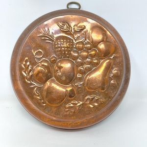May include: Round copper kitchen mold with a detailed embossed design of various fruits, including grapes, pears, and apples. The mold has a hanging loop at the top and is likely used for decorative or culinary purposes.