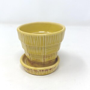 May include: A small, yellow ceramic planter with a matching saucer. The planter has a textured, basket-weave design. The saucer is round with a slightly raised edge. The planter and saucer are both a solid, pale yellow color.