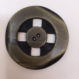 May include: A large, round, gold-colored button with a black center and four cutouts. The button has a raised, concentric circle design and two small holes.