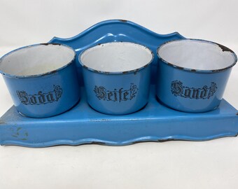 May include: A blue enamelware countertop dish rack with three small bowls. The bowls are labeled "Soda", "Seife", and "Sand".