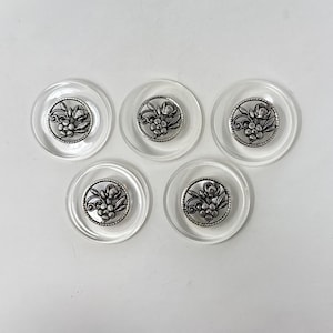 May include: Five clear plastic buttons with silver floral designs. The buttons are round and have a raised edge.