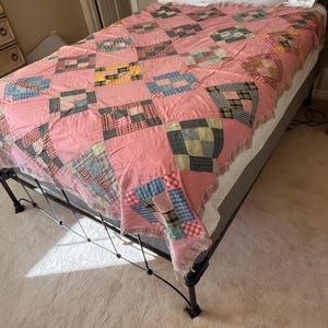 May include: A handmade quilt with a pink background and patchwork squares in various colors and patterns, including blue, red, and green. The quilt is draped over a bed with white pillows.