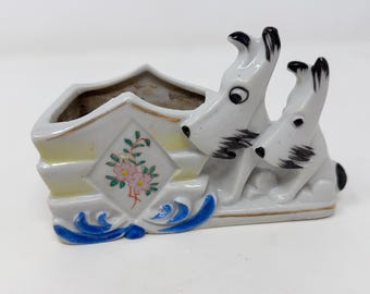 May include: A white ceramic planter with two dog figures. The planter has a diamond-shaped floral design and blue accents. The dogs have black markings and are sitting side-by-side.