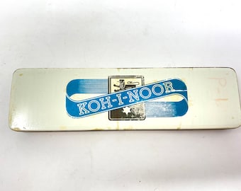 May include: A white rectangular tin box with a blue and white ribbon design. The ribbon has the text "KOH-I-NOOR" in white letters. The box has a lion crest with a star in the center.