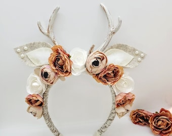 May include: A white headband with brown antlers and a floral crown of white and peach-colored flowers. The headband is decorated with small white pearls.