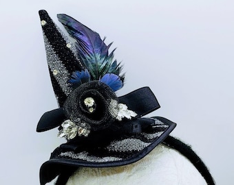 May include: Black witch hat headband with a black and silver patterned fabric, a black flower with rhinestones, and blue and black feathers.