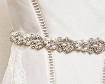 May include: A decorative belt featuring a repeating pattern of silver-toned metal filigree, clear faceted crystals, and small white faux pearls. The belt is worn around the waist of a white sleeveless dress with vertical lace trim detailing.
