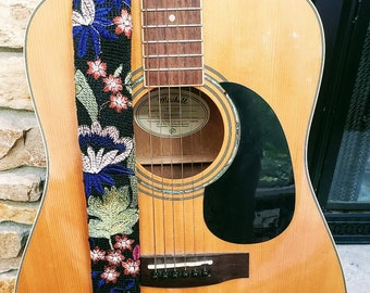 May include: A brown acoustic guitar with a black pickguard and a floral guitar strap. The strap is black with embroidered flowers in shades of blue, pink, and green. The strap is labeled "Ernie Ball".