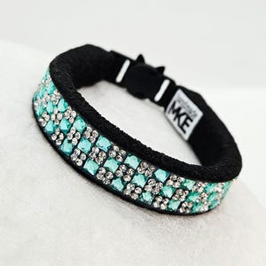 May include: Black hair tie with two rows of turquoise and clear rhinestones. The hair tie has a black band and a small tag with the letters "MKE". The rhinestones are square-cut and arranged in a grid pattern.