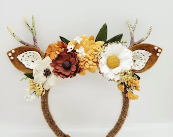 Puede incluir: Una diadema con cuernos de ciervo y orejas de fieltro marrón, adornada con una variedad de flores en tonos blancos, naranjas y amarillos. La diadema está envuelta en un material marrón texturizado, creando un aspecto rústico y natural.