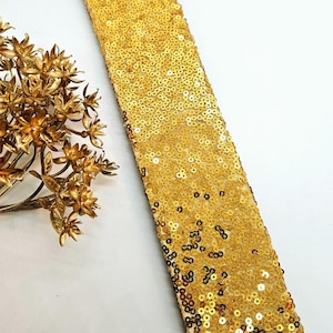 May include: A gold sequin trim, likely for embellishing garments or crafts, is displayed against a white background. The trim is densely covered in small, shiny sequins. A gold-colored floral decorative element is in the image.