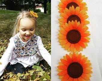 Puede incluir: Cuatro pinzas para el pelo de girasol de fieltro en tonos naranja y amarillo. Las pinzas están apiladas, con el girasol más grande en la parte superior. Los girasoles tienen centros marrones y están rodeados de pétalos amarillos.