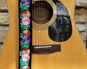 May include: A black guitar strap with a colorful floral pattern. The strap is embroidered with pink, red, green, and purple flowers. The strap is attached to a brown acoustic guitar. The strap has the text "Ernie Ball" in gold lettering.