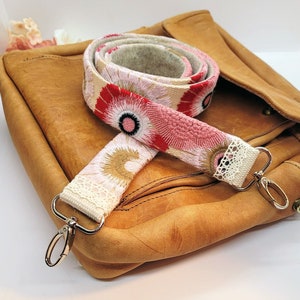 May include: A close-up of a pink and red floral patterned fabric bag strap with white lace trim and silver clasps. The strap is on top of a brown leather bag.