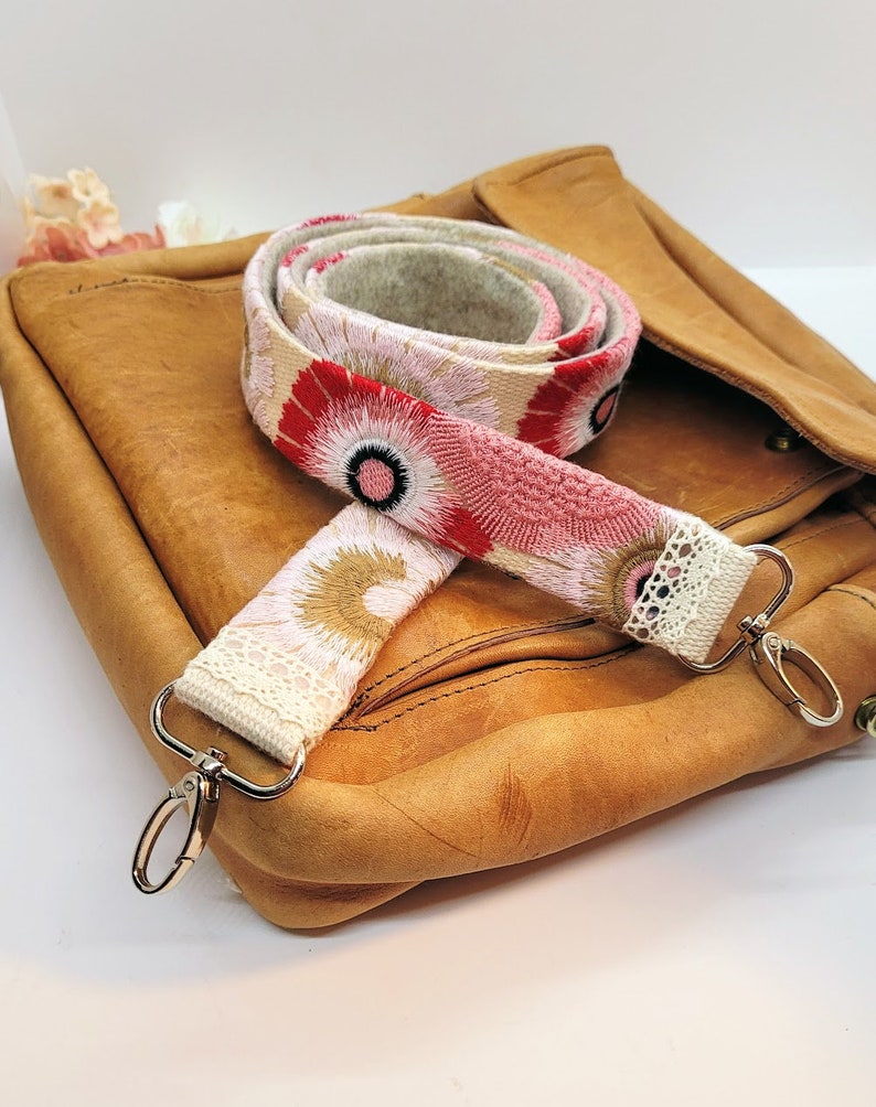 May include: A close-up of a pink and red floral patterned fabric bag strap with white lace trim and silver clasps. The strap is on top of a brown leather bag.