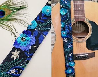 May include: A black guitar strap with blue and green embroidered flowers and sequins. The strap is attached to a brown acoustic guitar. The strap has the text "Fender Roll" on it.