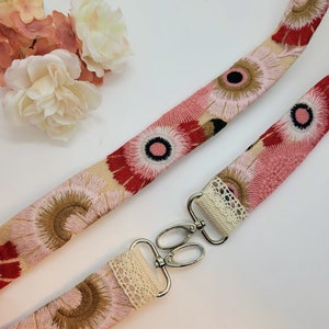 May include: A pink and red floral patterned fabric strap with a white lace trim and silver metal clasps. The strap is designed for a bag or purse.