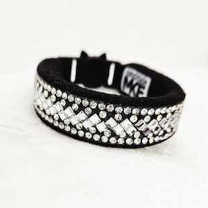 May include: Black headband with a sparkling, geometric pattern of clear rhinestones. The band is made of black fabric and features a small tag with the letters "MKE". The design is symmetrical and eye-catching.