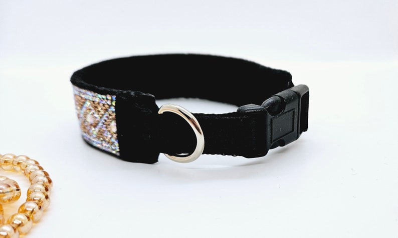 Puede incluir: Un collar de perro negro con un anillo en D plateado y una hebilla de pl&aacute;stico negro. El collar tiene una banda decorativa de lentejuelas y cuentas iridiscentes.