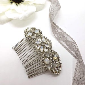 May include: A silver-toned hair comb with an ornate design featuring clear and iridescent crystals. The comb has a series of thin, evenly spaced tines. A white flower and gray lace ribbon are in the background.
