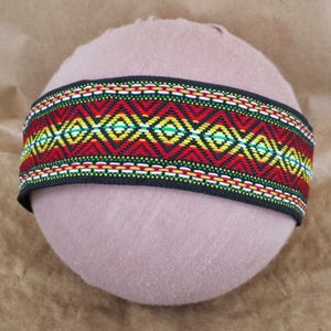 May include: A black woven band with a colorful geometric pattern in red, yellow, green, and white. The band is on a light brown surface.