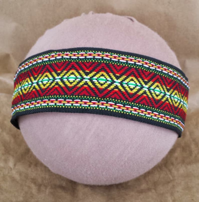 May include: A black woven band with a colorful geometric pattern in red, yellow, green, and white. The band is on a light brown surface.
