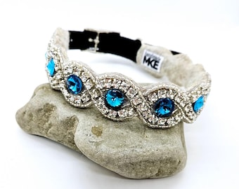 May include: A silver and white dog collar with a row of blue rhinestones. The collar has a black buckle and a white fabric lining. The collar is on a gray rock.