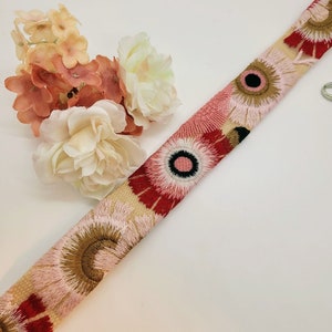 May include: A beige fabric ribbon with embroidered pink, red, and brown flowers. The flowers have a black center with a white circle inside.
