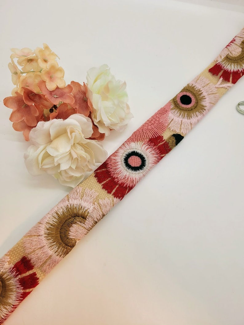 May include: A beige fabric ribbon with embroidered pink, red, and brown flowers. The flowers have a black center with a white circle inside.
