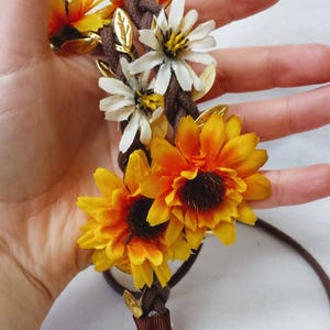 May include: A brown braided headband with yellow and white fabric flowers. The headband has gold leaf accents and a brown ribbon tie.