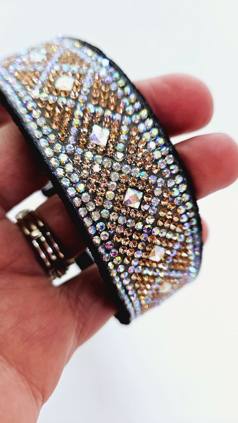 Puede incluir: Un brazalete de cuero negro con un patr&oacute;n geom&eacute;trico de diamantes de imitaci&oacute;n iridiscentes y dorados.