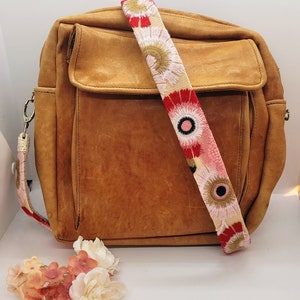 May include: A brown leather satchel bag with a long, colorful strap featuring embroidered flowers in shades of red, pink, and white.
