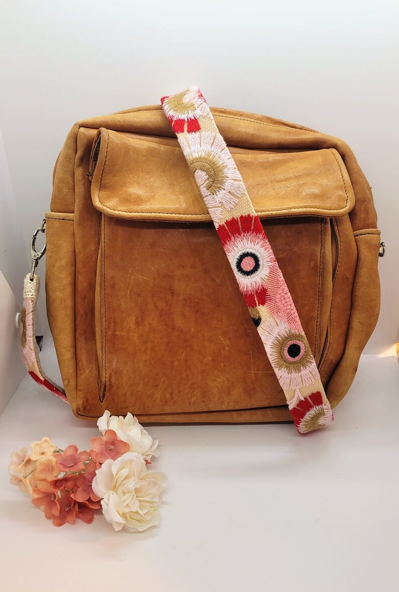 May include: A brown leather satchel bag with a long, colorful strap featuring embroidered flowers in shades of red, pink, and white.