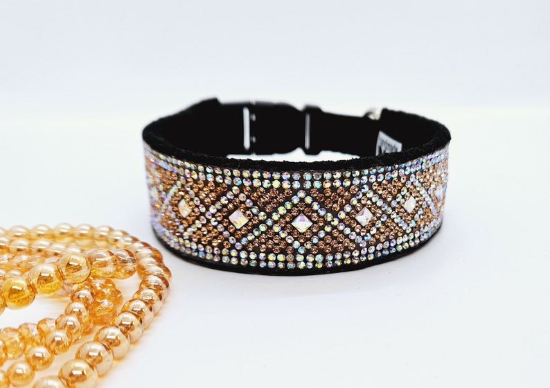 Puede incluir: Un collar de perro negro con una banda ancha de diamantes de imitaci&oacute;n iridiscentes en un patr&oacute;n geom&eacute;trico. Los diamantes de imitaci&oacute;n son en tonos dorados y transparentes.