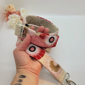 May include: A pink and red floral patterned fabric strap with a white lace trim and a silver metal key ring. The strap is folded in half and held in a hand.